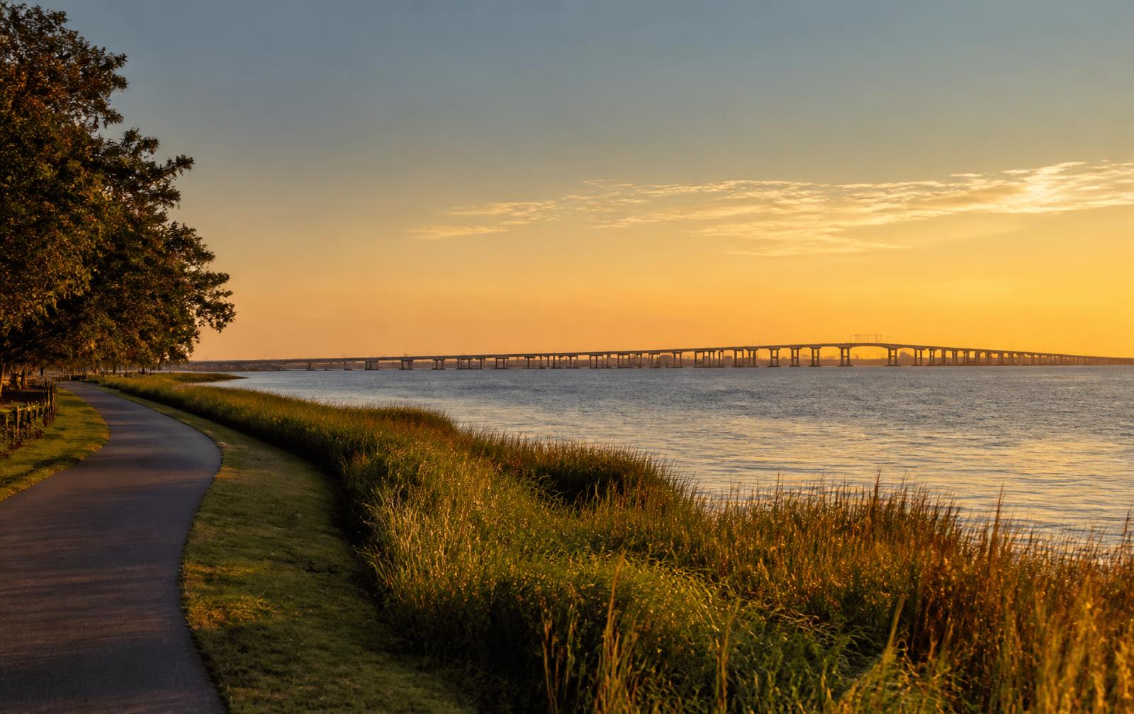 Waterfront path along the Hampton Roads bay at sunset with a long bridge in the distance